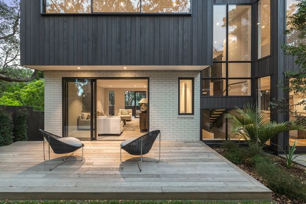 Exterior view of a contemporary multi-level home with dark vertical cladding, pale brickwork, extensive black-framed glazing, and a broad timber deck. Two outdoor lounge chairs occupy the deck, while the glazed ground floor reveals a living area and an internal stair. Landscaped planting borders the house.