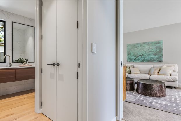 Interior view from a hallway or entry area looking toward an open bathroom on the left and a living room on the right. The bathroom features a floating dark wood vanity, white tiled walls, a large mirror, and a glass shower enclosure. The living room contains a light sofa, textured round coffee tables, a pale rug, and a green abstract artwork. Light timber flooring and white walls create a bright, contemporary interior.