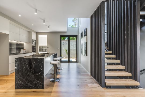 Contemporary open-plan kitchen and circulation space with white full-height cabinetry, a dark marble island with integrated seating, timber flooring, and a sculptural open-riser staircase screened by vertical dark timber slats. The room is bright and refined, with glazed doors opening to a small outdoor area and a high clerestory-like window above the hallway.