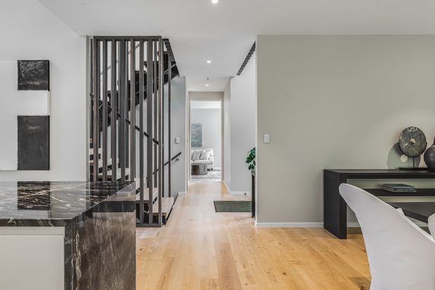 Contemporary open-plan interior showing a kitchen island with dark veined stone surfaces, a timber staircase enclosed by vertical dark metal battens, pale timber flooring, and a compact dining or study area with a black built-in desk. A hallway leads toward a softly furnished bedroom in the background.