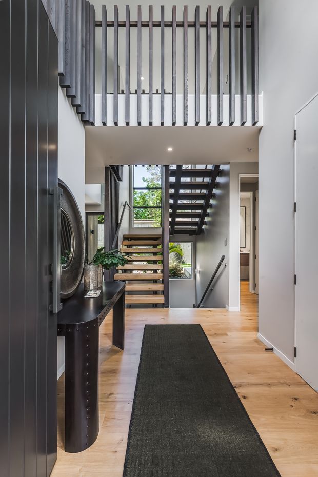 A contemporary residential entry hall with a timber floor, dark built-in cabinetry and console, a black runner, and an open-riser staircase leading to a mezzanine or upper landing. Vertical dark timber balustrade slats create a strong architectural feature above the stair void, while tall glazing brings in natural light and views of greenery. The space has white walls, concealed lighting, and a refined monochrome-and-timber palette.