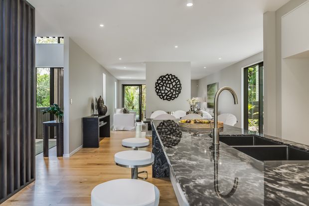 A bright, open-plan contemporary interior viewed from the kitchen island. The space features a large dark veined stone island with integrated double sink and curved brass-toned tap, white round stools, light timber flooring, white walls and cabinetry, recessed ceiling lights, and black-framed glazing. A dining and living area extends beyond the kitchen, with sculptural wall art and greenery visible through the windows.