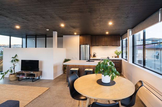 Open-plan contemporary apartment interior combining a compact kitchen and dining area with a partially separated living room. The space features dark timber-lined ceilings, warm timber kitchen cabinetry, a light tiled backsplash, large black-framed windows, polished concrete flooring, and a round timber dining table. A low white partition defines the living zone, which contains a television and understated furniture. The room is bright and inviting with urban views through the glazing.