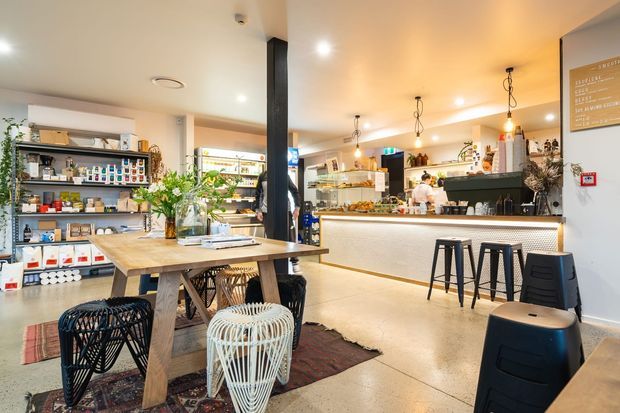 Bright contemporary café interior with a casual dining area in the foreground and a service counter, espresso machine, display shelving, and food preparation area at the rear. The space features polished concrete flooring, warm timber tables, black structural columns and stools, pendant lighting, open retail shelving, and a welcoming commercial fit-out.