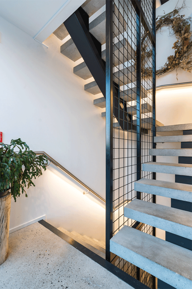 A contemporary interior stairwell featuring a floating concrete staircase with dark metal framing and a black grid-style balustrade. The stairs connect multiple levels around a central void, with integrated warm linear lighting, pale walls, and a terrazzo-like light floor. A large indoor plant and dried botanical feature add organic detail.