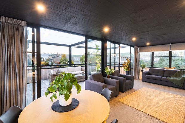 A spacious contemporary living room with floor-to-ceiling black-framed glazing opening onto a balcony or terrace. The room features a large round timber dining table, upholstered armchairs, a dark sectional sofa, indoor plants, textured curtains, and a broad natural-fibre rug. A dark timber-toned ceiling with recessed downlights creates a warm, refined interior, while the glazing provides abundant daylight and views of the surrounding urban neighbourhood.
