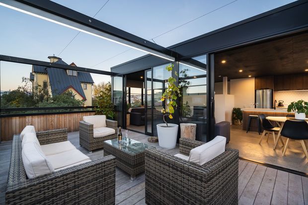 A contemporary indoor-outdoor living area with a furnished timber-deck terrace opening directly into a modern kitchen. The terrace has woven outdoor lounge seating, a glass coffee table, planter, and timber privacy balustrade beneath a dark exposed steel pergola with linear lighting. Full-height black-framed glazing connects the terrace to the kitchen, which features dark timber cabinetry, a light island benchtop, and warm recessed lighting. A neighbouring pitched-roof house is visible beyond the terrace.