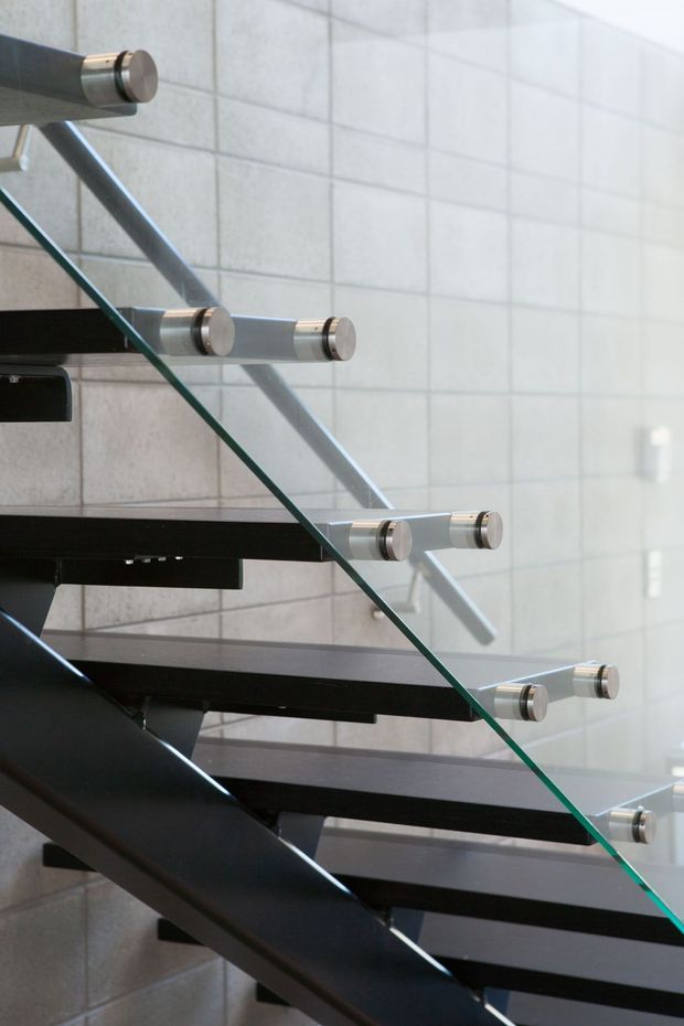 Close-up architectural photograph of a modern floating staircase with dark timber or metal treads, a substantial dark structural stringer, and a clear glass balustrade secured by cylindrical brushed-metal fittings. The staircase is set against a light grey tiled wall, with strong diagonal lines and abundant natural light creating a clean contemporary feel.