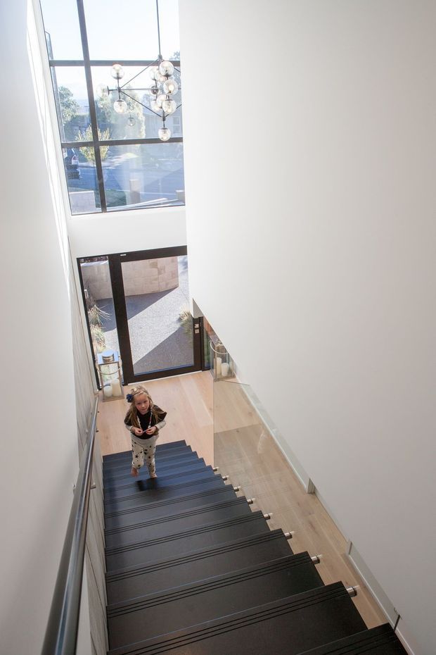 Bright contemporary stairway viewed from above, with broad dark floating stair treads, pale timber flooring, white walls, a glass balustrade, and a tall black-framed glazed entry facade. A sculptural cluster pendant hangs within the double-height stair void, with daylight entering through the large windows.