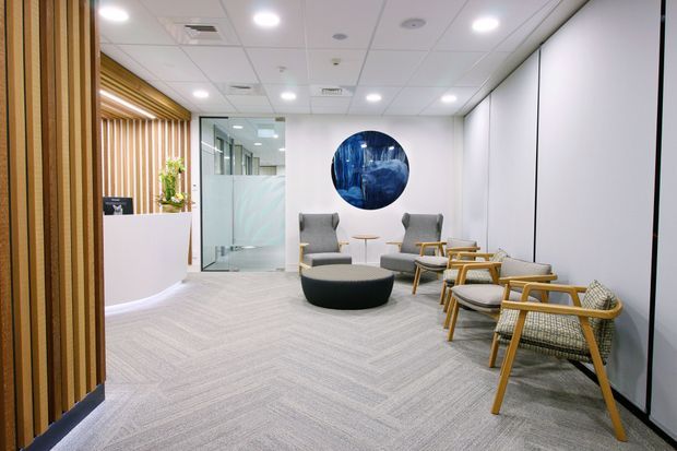 A bright, contemporary commercial reception and waiting area with a curved white reception desk, timber slat feature wall, glass-partitioned room, carpeted floor, and a cluster of upholstered lounge chairs around a round ottoman and small table. A large circular blue artwork provides a focal point on the white wall.