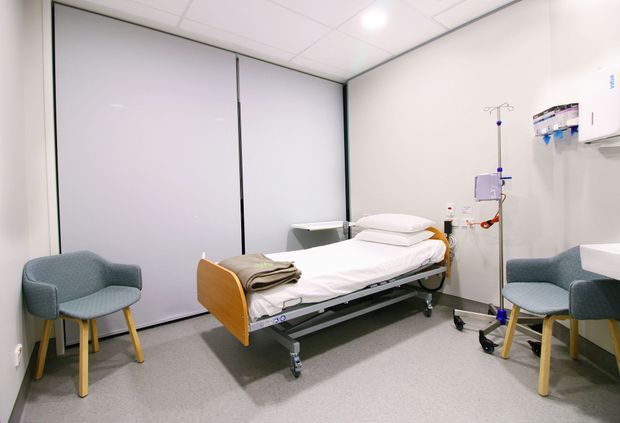 A clean, modern clinical patient room with a hospital bed centered between two upholstered visitor chairs. The room has pale walls, seamless light flooring, privacy roller blinds, a suspended medical equipment rail, IV stand, wall-mounted clinical supplies, and a white suspended ceiling with recessed lighting.