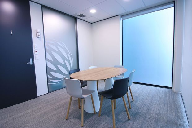 A compact contemporary meeting room with a round light-wood table surrounded by mixed white and dark upholstered chairs. The room has grey carpet, frosted glass partitions with a leaf-pattern privacy graphic, a large translucent window or screen, white acoustic ceiling tiles with recessed lighting, and a dark entry door.