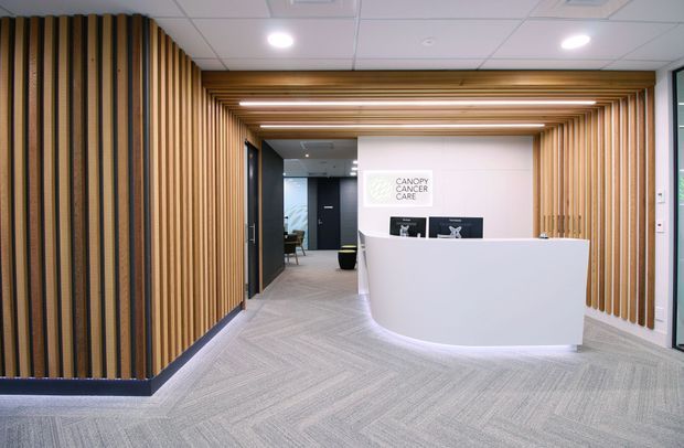 A contemporary commercial reception area with a curved white reception desk, integrated display screens, and a visible Canopy Cancer Care sign. The space features extensive vertical timber slat wall and ceiling detailing, recessed linear lighting, grey patterned carpet, dark door surrounds, and a suspended acoustic tile ceiling. An adjoining office or waiting area is visible beyond the reception zone.