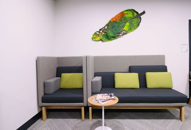 A contemporary commercial lounge or waiting area with two large grey upholstered booth-style sofas, lime green and dark cushions, a small round timber-topped side table, and a colourful wall-mounted artwork. The space has pale walls, grey carpet, and simple modern furnishings.