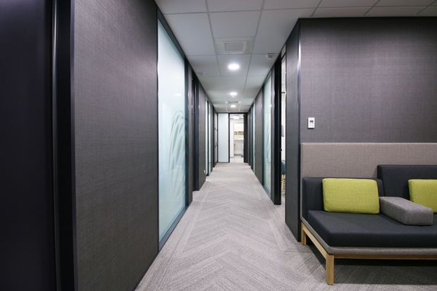 A modern commercial office corridor with dark textured wall panels, full-height glazed office partitions, a suspended acoustic tile ceiling, and light grey patterned carpet. A contemporary black sofa with lime green and grey cushions sits in a small waiting area along the right wall.