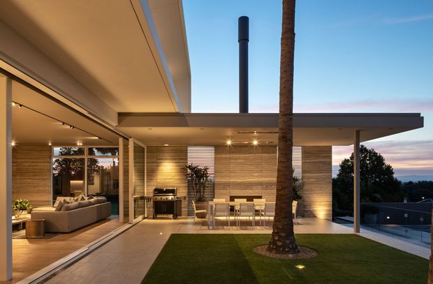 Contemporary flat-roofed residential home photographed at dusk, showing a covered outdoor entertaining terrace with a built-in barbecue, dining table, tiled paving, lawn and a palm tree. Floor-to-ceiling glazing reveals an adjacent living room with a large sectional sofa. The house features clean horizontal lines, warm recessed lighting, pale textured wall cladding and slender dark structural posts.