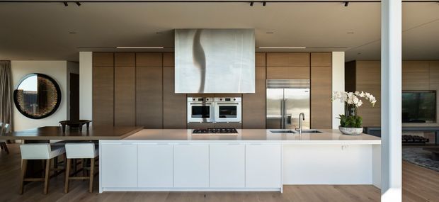 Contemporary open-plan kitchen with a large white island, integrated cabinetry, timber wall panels, built-in ovens, stainless steel refrigerator, and a sculptural pale stone or plaster rangehood feature. A dining area and living space are visible beyond, with warm timber flooring and restrained neutral finishes.