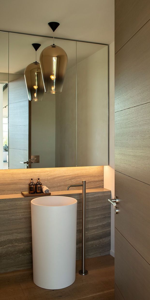 A refined contemporary bathroom featuring a freestanding cylindrical white basin, tall brushed-metal tapware, large mirrored wall panels, warm stone-look wall surfaces, timber flooring, and sculptural amber pendant lights. A textured timber door and matching vertical wall panelling frame the space, creating a warm, softly lit atmosphere.