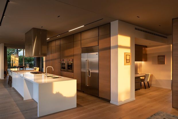 Warm contemporary open-plan kitchen and dining area with a large white island, integrated stainless-steel appliances, extensive dark timber cabinetry, timber flooring, and a compact dining nook. Late afternoon sunlight creates strong golden highlights across the cabinetry and walls.