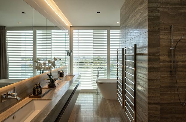A sophisticated contemporary bathroom with a long double vanity and illuminated mirror wall on the left, a freestanding bathtub positioned in front of large windows with horizontal blinds, and a walk-in shower clad in dark, horizontally textured stone-look panels on the right. A heated towel rail, recessed ceiling lights, timber-toned vanity surfaces, and warm integrated lighting create a refined spa-like atmosphere.