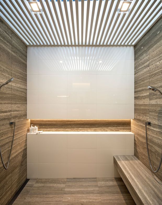 Contemporary bathroom with a spacious walk-in shower, large-format glossy white wall tiles, warm brown stone-look wall and floor tiles, and a built-in tiled bench. A dramatic white slatted ceiling with recessed lighting creates a strong linear feature, while handheld shower fittings are mounted on both side walls.