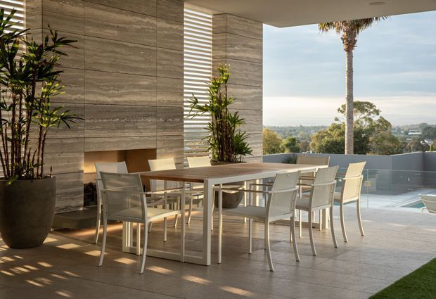 A refined outdoor dining terrace with a large rectangular table and contemporary chairs, sheltered beneath a flat-roofed canopy. The space features large-format grey stone or concrete-look wall and floor finishes, a linear fireplace, vertical screening, potted tropical plants, and views toward a landscaped suburban setting. A swimming pool edge is visible at the right.