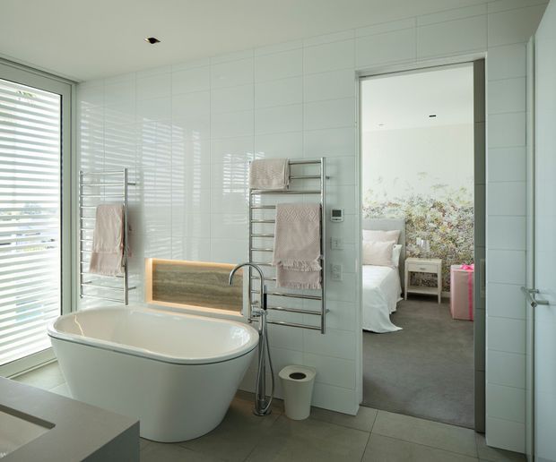 Bright contemporary bathroom with a freestanding white bathtub, wall-mounted towel warmers, white tiled walls, grey floor tiles, and a timber-toned illuminated bath niche. A bedroom is visible through the open doorway, with floral wallpaper and a neutral upholstered bed.