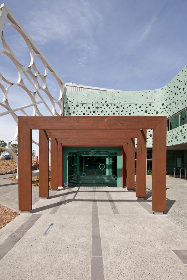 Exterior view of a contemporary commercial building entrance. A broad concrete forecourt with dark linear inlays leads through a sequence of substantial timber portal frames to glazed entry doors. The surrounding facade features pale green perforated panels with varied circular openings and a dramatic curved form, while a large white sculptural screen with overlapping circular motifs rises along the left side. The image is architectural and strongly geometric, photographed in bright daylight.