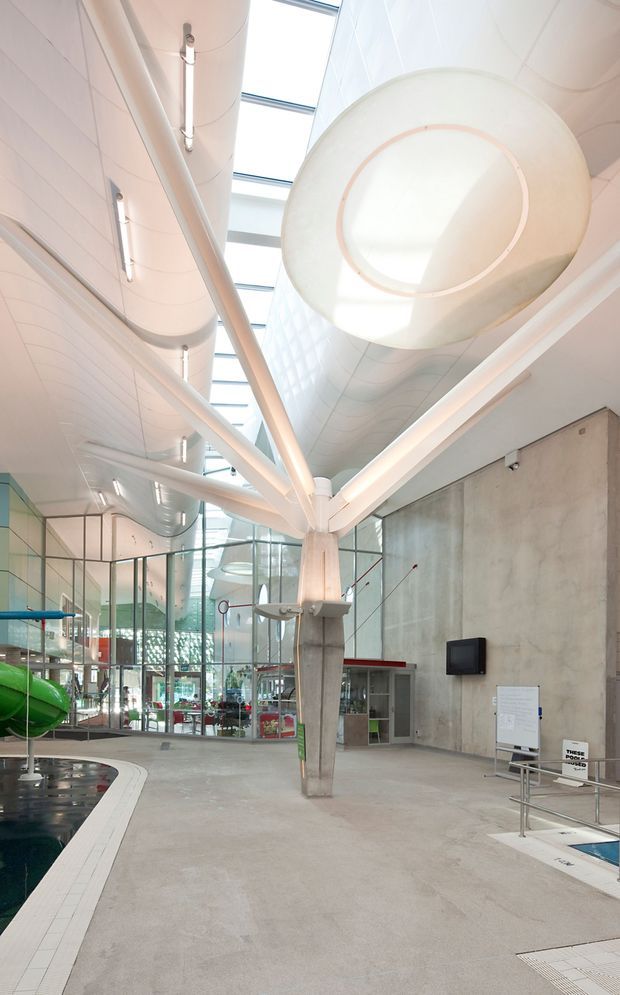 A large, bright indoor aquatic facility with a pool edge in the foreground, expansive glass walls, skylights, exposed concrete, and a dramatic white sculptural suspended ceiling or ventilation element. The space has a high ceiling, curved architectural forms, tubular services, and contemporary institutional detailing.