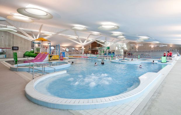A large modern indoor aquatic centre with multiple swimming pools, a shallow children's play pool with colourful slides and water features, lap lanes, tiled surrounds, and distinctive white branching structural roof supports. The bright space is illuminated by circular ceiling lights and skylight-like fixtures.