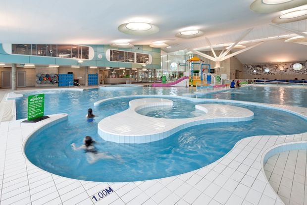 Bright indoor aquatic centre with multiple interconnected swimming pools, curved white-tiled pool edges, a shallow leisure pool, waterslide and colourful children's play structure. The large glazed and open interior has a high sculptural ceiling with circular skylight-like light fixtures, upper-level viewing areas, and changing or service facilities around the perimeter.