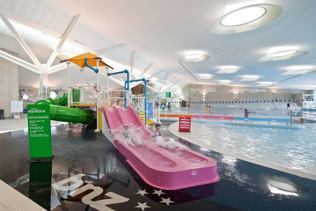 Interior view of a large modern aquatic centre with multiple swimming pools, waterslides, colourful play structures, and a high white vaulted ceiling. The glossy dark floor reflects large oval ceiling lights, while pool safety signage and lane markings are visible throughout the bright recreational facility.
