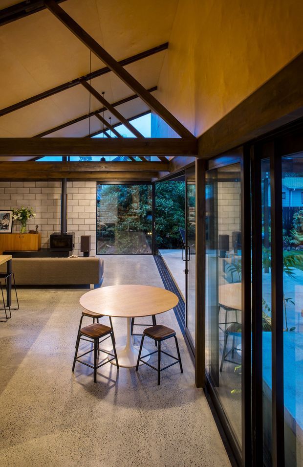 A warm, open-plan contemporary interior with a dining area in the foreground and a living area beyond. The space features exposed dark timber roof beams and rafters, a high pitched ceiling, polished concrete flooring, pale concrete block walls, extensive black-framed glazing, and a strong indoor-outdoor connection to a landscaped garden and pool visible through the glass. A round timber dining table with stools sits beside the glazed wall, while a sofa, compact fireplace, and low timber cabinetry occupy the adjoining living zone.