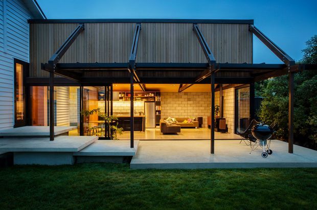Twilight view of a contemporary house extension opening onto a broad concrete terrace and lawn. The covered outdoor area features a timber-clad, steel-supported canopy with exposed diagonal bracing, full-height glazing, and an illuminated open-plan interior visible beyond. A built-in-style bar or kitchen area and lounge seating are visible inside, while a barbecue stands on the terrace.