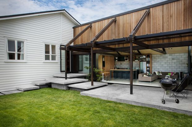 A contemporary single-storey home extension opens onto a covered outdoor entertaining area. The original white weatherboard house is joined to a timber-clad addition with dark steel structural beams, a concrete terrace, and extensive glazing. Through the open facade, a compact kitchen and living area are visible, while the foreground shows a lawn and barbecue.