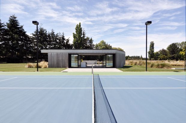 A modern, low-profile pavilion positioned behind a blue outdoor tennis court. The building has dark vertical cladding, a shallow flat roof, and a broad glazed opening revealing a compact interior. Tall black court lights frame the scene, with landscaped grass and mature trees surrounding the facility.
