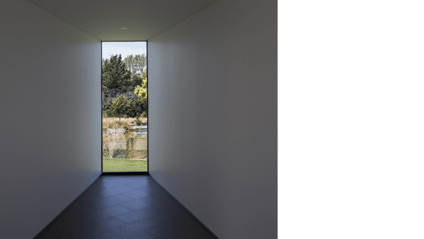 A narrow, minimalist interior passage or entry corridor with smooth dark grey walls and ceiling, a dark tiled floor, and a tall, slender glazed opening at the far end framing landscaped greenery outside. The restrained palette and strong linear perspective create a quiet, contemplative atmosphere.