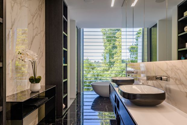 Luxurious contemporary bathroom with a double vanity, two vessel basins, dark built-in cabinetry, pale marble-look wall surfaces, and a freestanding bathtub. Full-height glazing with horizontal screening brings in views of leafy greenery, while glossy dark stone-look flooring creates strong reflections.