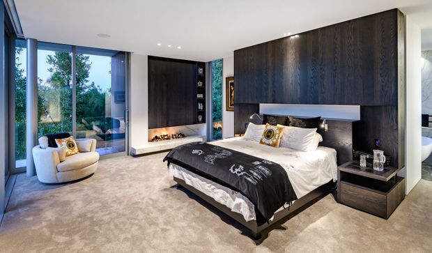 Luxurious contemporary bedroom with a large upholstered bed, dark timber feature wall and integrated bedside joinery, a linear fireplace, floor-to-ceiling glazing, and a soft neutral carpet. The room opens toward a leafy outlook and includes a marble-lined bathroom glimpse at right.