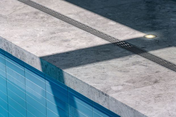 Close-up architectural detail of a swimming pool edge, showing blue tiled pool walls, a pale grey stone or concrete deck, a linear drainage channel, and a small recessed deck light.