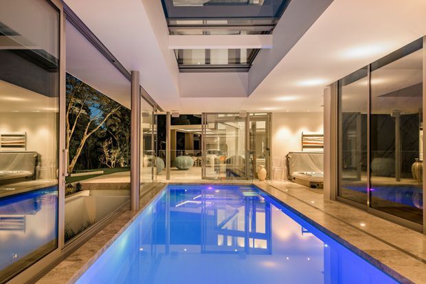 A luxurious indoor swimming pool extending through a bright, glass-lined contemporary interior. Floor-to-ceiling glazing, reflective stone or tile flooring, warm recessed lighting, and a dramatic overhead skylight create a seamless connection to the landscaped exterior. Furnished living areas and decorative objects are visible beyond the pool.