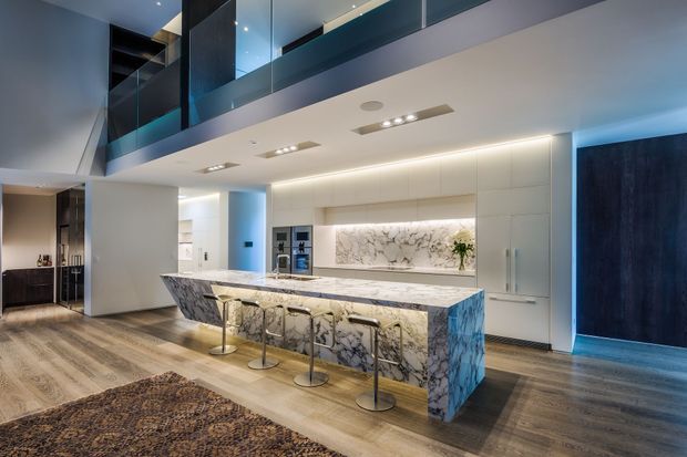 A sophisticated open-plan kitchen beneath a mezzanine or upper-level void. The space features a large waterfall-edge marble or heavily veined natural-stone island with integrated seating, white built-in cabinetry, a matching illuminated stone splashback, timber-look flooring, recessed and linear lighting, and dark blue-black architectural joinery and doors. Glass balustrading and upper-level glazing contribute to the contemporary, high-end feel.
