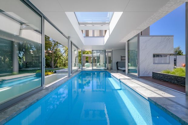 A bright contemporary indoor-outdoor pool courtyard with a long rectangular swimming pool running through a glass-lined central space. Floor-to-ceiling glazing, white geometric volumes, a large skylight, pale stone paving, and landscaped edges create a clean, open architectural composition.
