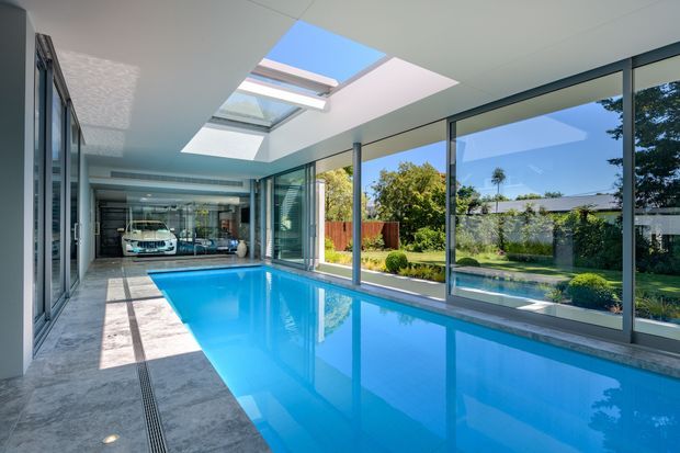 Bright enclosed indoor swimming pool with large floor-to-ceiling glazed walls, a skylight, pale stone-look flooring, and a minimalist white ceiling. The pool room opens visually toward a landscaped garden, with a garage and car visible in the background.