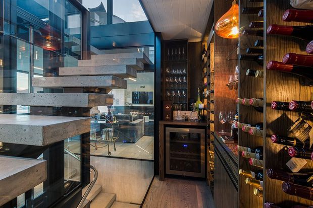 A sophisticated wine storage and serving area beside a sculptural floating concrete staircase. The compact space features dark timber wine racks, extensive bottle storage, glassware shelving, a built-in wine refrigerator, warm pendant lighting, and glass partitions overlooking a contemporary interior. The combination of concrete, dark wood, glass, and warm lighting creates a luxurious, dramatic atmosphere.