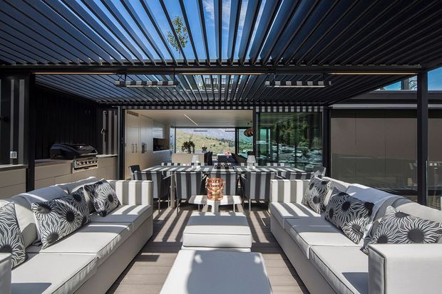 A contemporary covered outdoor entertaining area with a large louvred pergola, two white sectional sofas, patterned cushions, and a central ottoman. A built-in outdoor kitchen with grill and cabinetry sits to the left, while a bar-height island with stools connects the space to a glazed interior kitchen or living area. Strong linear shadows fall across the pale flooring and furnishings, creating a bright, refined indoor-outdoor setting.