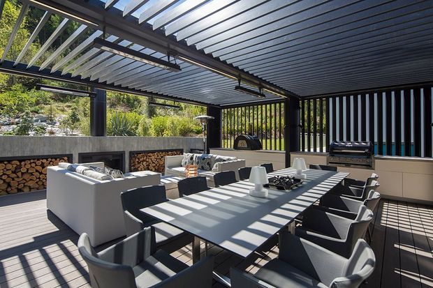 A sophisticated covered outdoor entertaining area with a large dining table, upholstered dining chairs, lounge seating, a built-in barbecue, firewood storage, and a compact outdoor fireplace or pizza oven. The space features a dark metal pergola and slatted roof with strong linear shadows, black structural posts and screens, pale cabinetry, and a timber-look deck. Lush landscaped planting and a rocky garden setting are visible beyond the open sides.