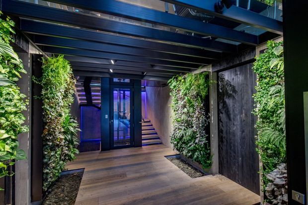 A dramatic covered entry passage with dark exposed steel beams, timber-look flooring, vertical living walls, and dark textured wall panels. The passage leads to glazed doors and a short stairway, with cool blue-purple lighting visible beyond. The design combines lush greenery with an industrial, high-contrast architectural fit-out.