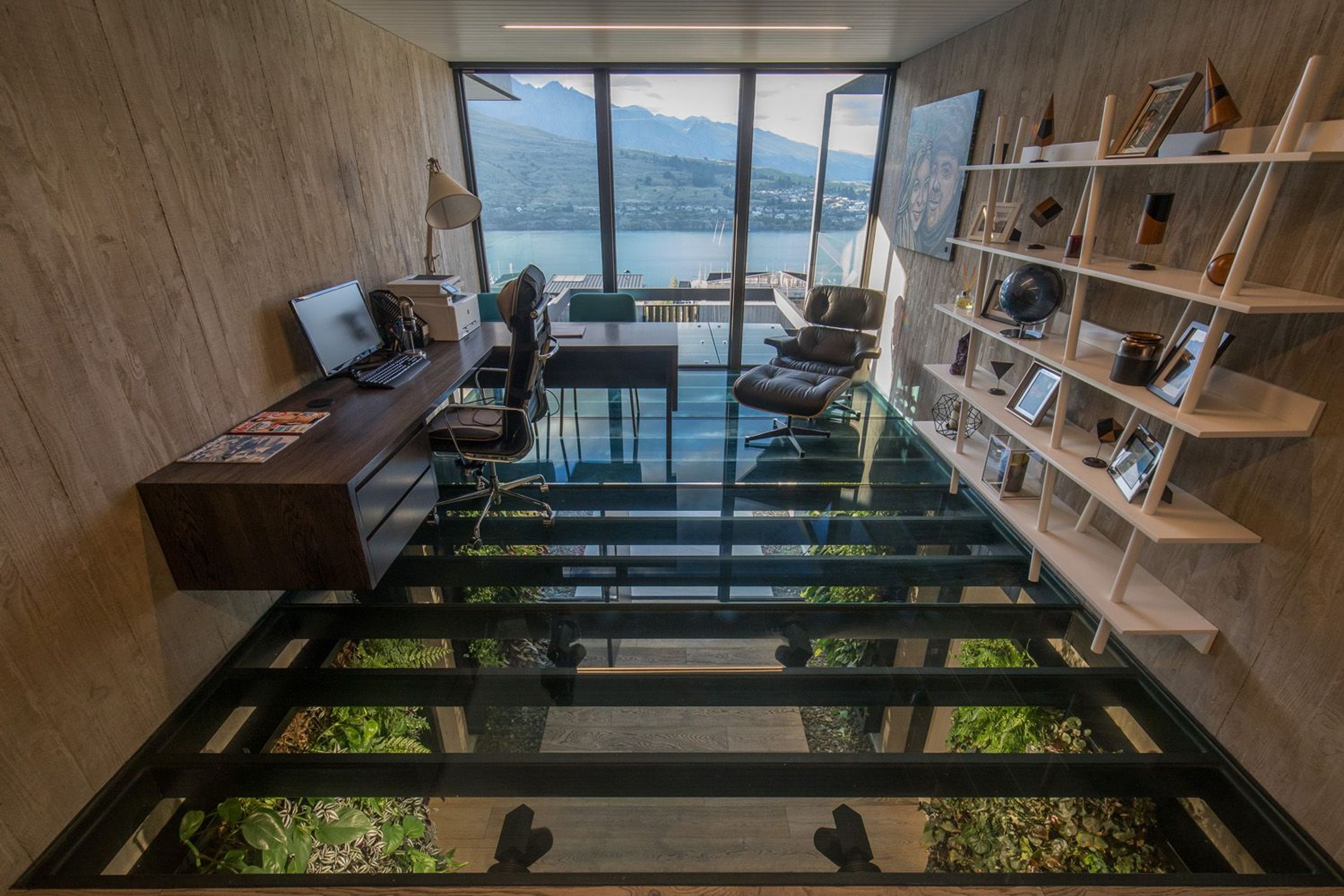 The glass floor of the office looks down into the entrance foyer, while floor-to-ceiling windows look out over the lake and mountains, framed by shuttered concrete walls.