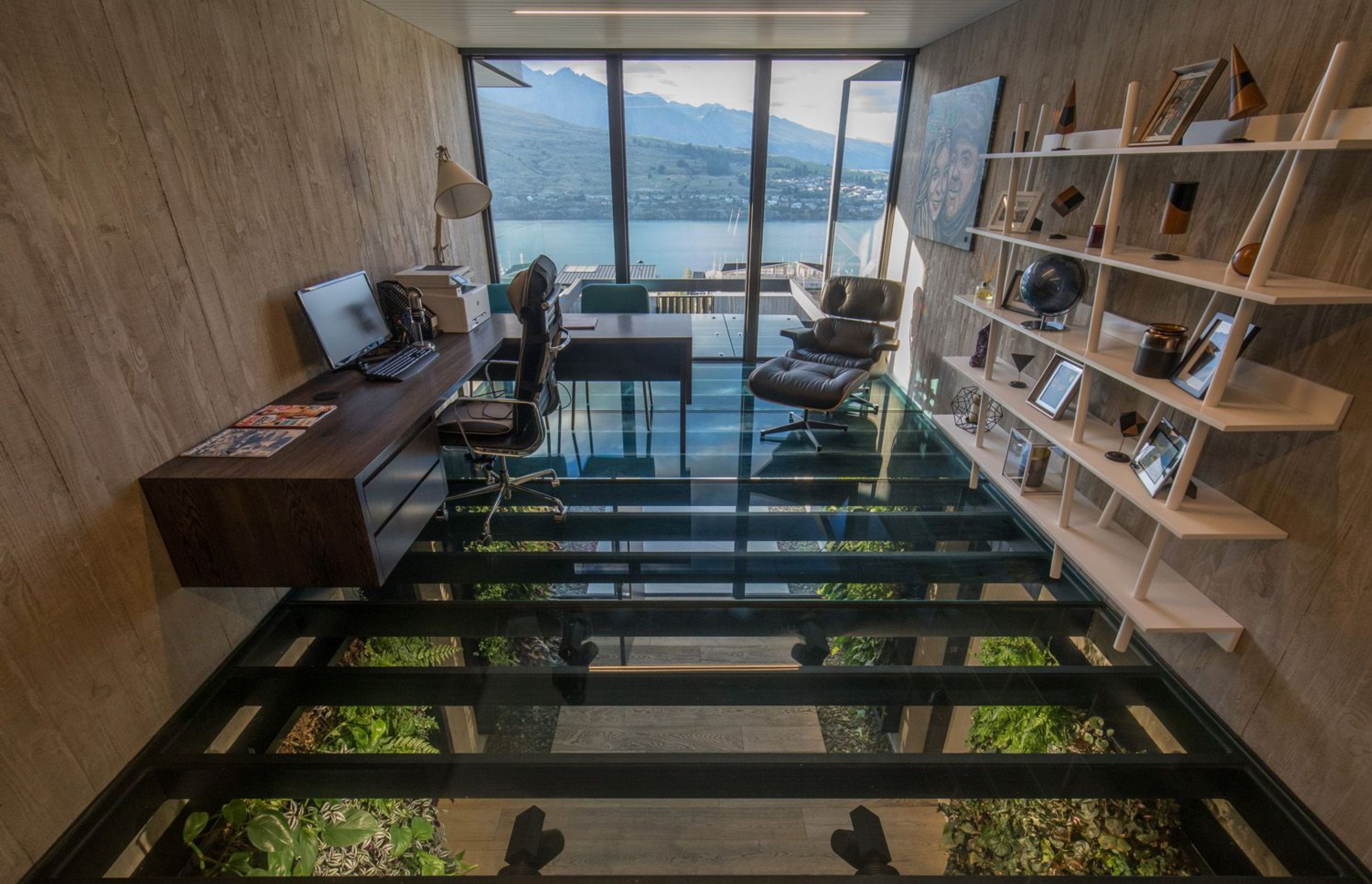 The glass floor of the office looks down into the entrance foyer, while floor-to-ceiling windows look out over the lake and mountains, framed by shuttered concrete walls.